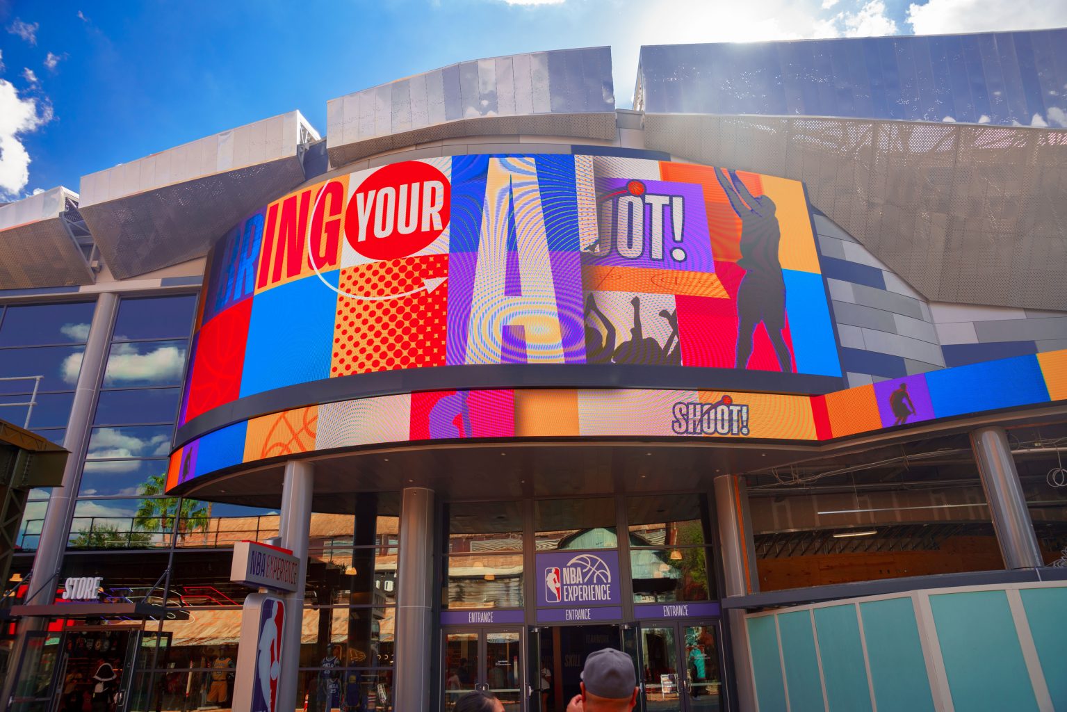 Storefront of the NBA store at DIsney Springs with a large, animated sign taking up most of the photo with a basketball player graphic and multi colored graphics and words. There are people at the bottom of the picture walking in the store, with a sign on the door that says "NBA Experience"