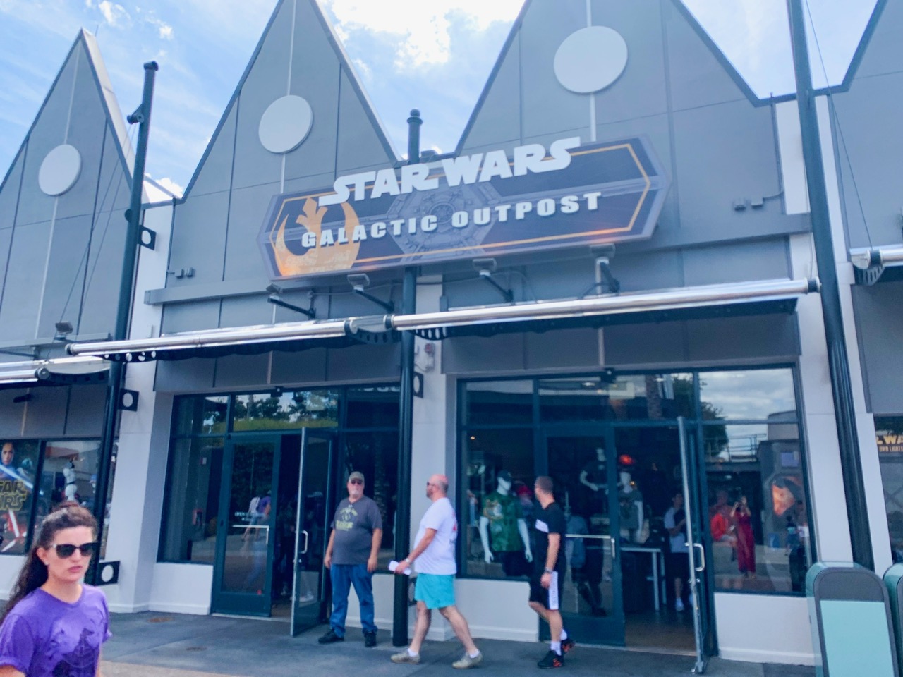 Storefront of the Star Wars Outpost which is a silver building with two large triangles on top with a sign that says "Star Wars Galactic Outpost" and people walking in and out of the store