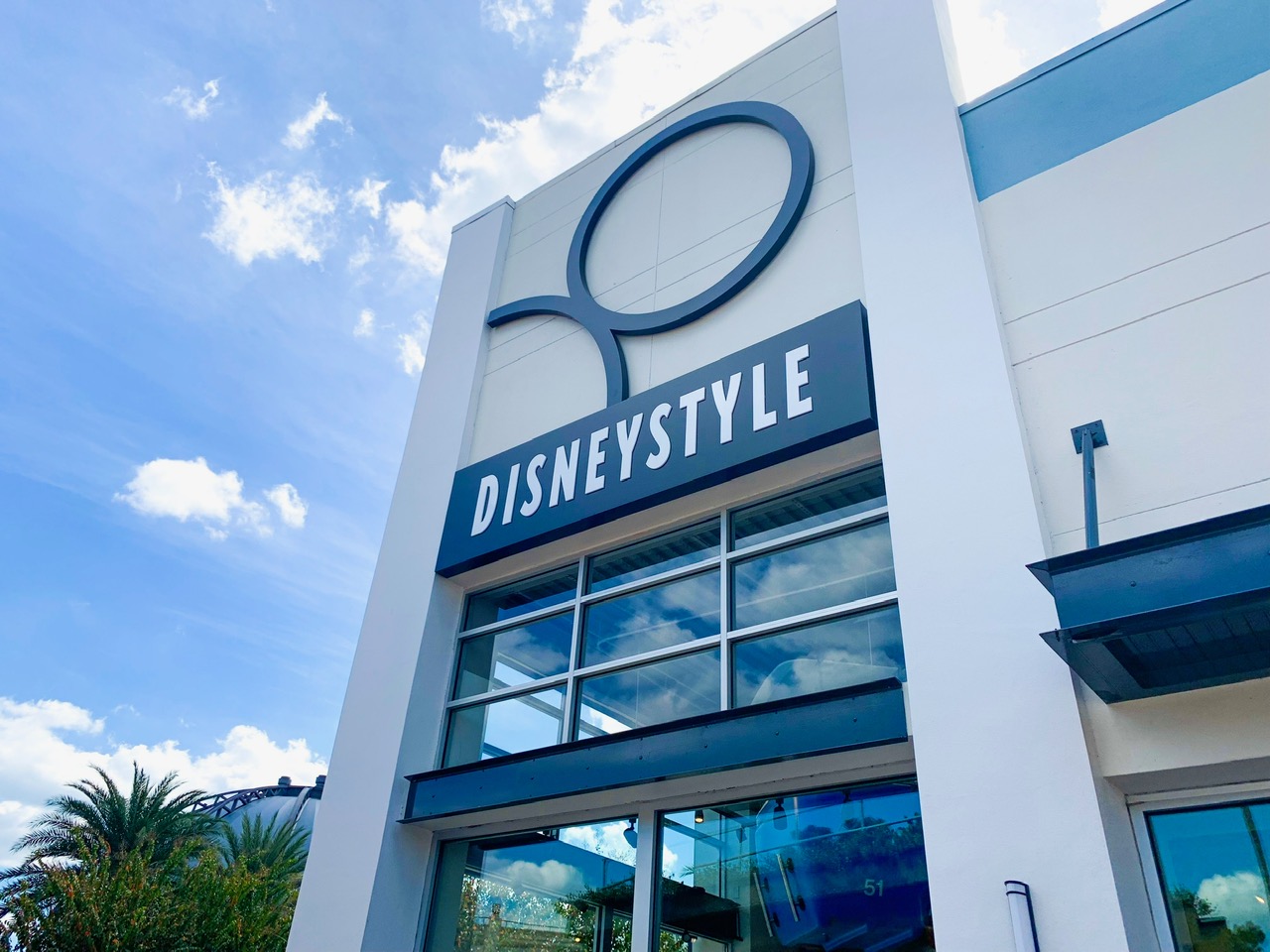 Storefront of DisneyStyle at Disney Springs. The storefront is an off white color with large windows and a dark blue sign with white, all capital DisneyStyle letters. There is a blue sky and clouds in the background.