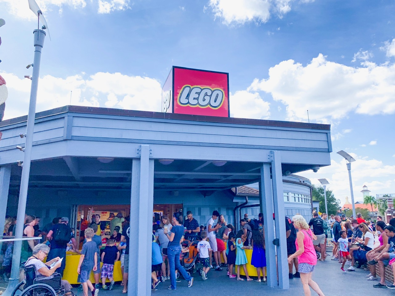 A crowd of people outside of the Lego store with white pillars and a red sign with white "lego" letters on top on a blue sky and cloudy day