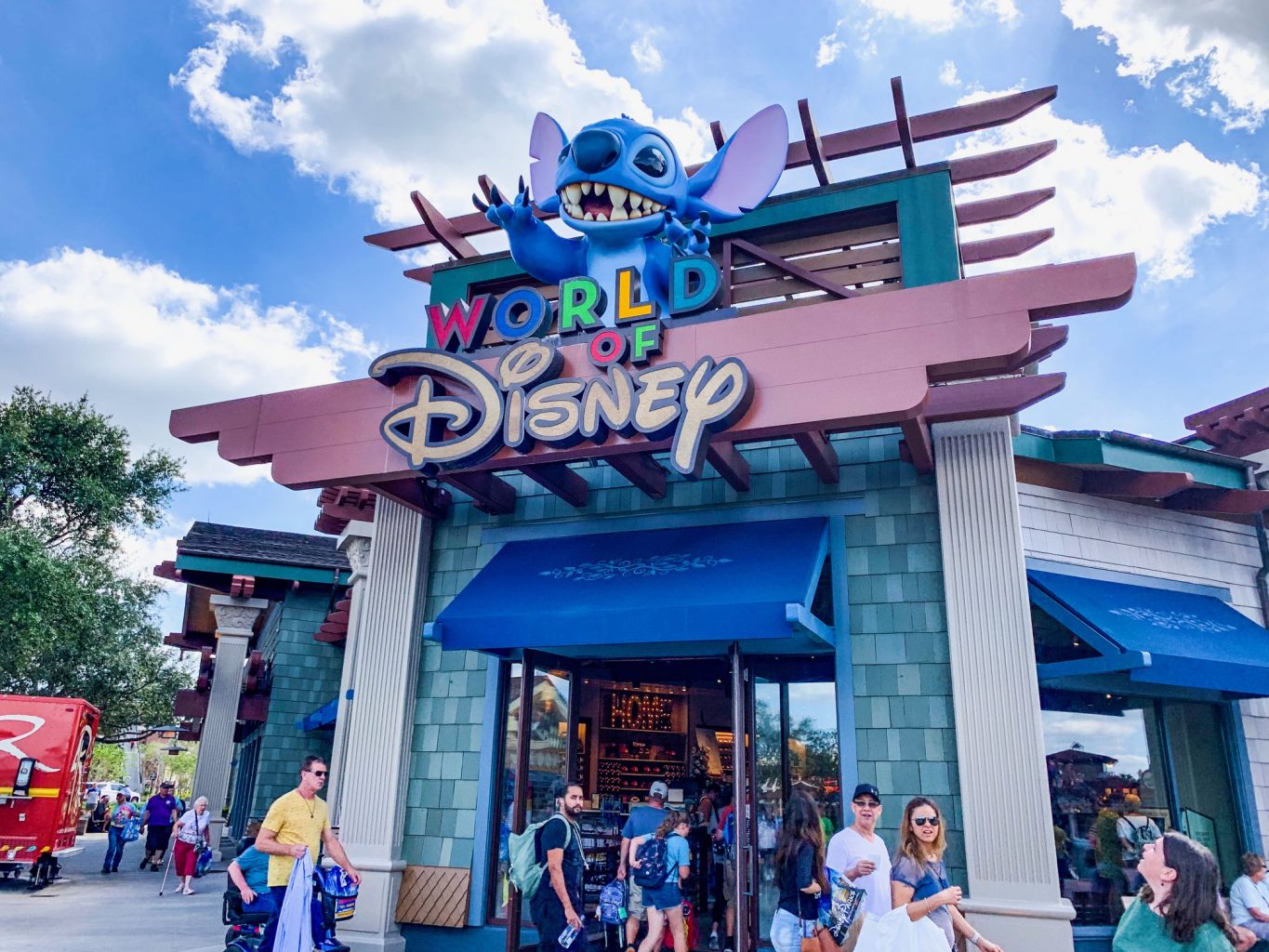 Storefront of World of Disney store at Disney Springs. It is a large grey store, with lots of people outside walking around, and has a blue awning. The store name is on a maroon background with multicolored letters for "World of" and then the Disney font in a soft yellow. 