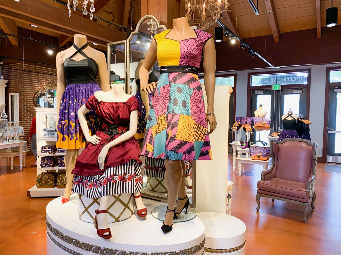 Set of three dresses on display inside the Marketplace Co Op store at Disney Springs. The dress on the far right has a color block yellow and pink top and a pink, yellow, and blue skirt with polka dots in some of the multi colored squares. The middle dress is dark red with a black and white striped under skirt peeking out from the bottom. The far left dress has a black criss-crossed halter top with a purple skirt with a flame pattern on the bottom. 