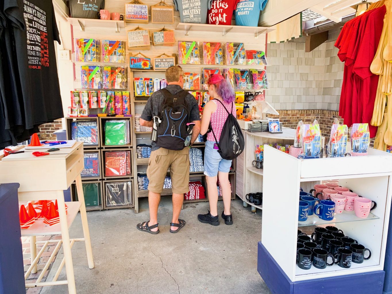 A couple shopping at the Left Hand Store at Disney Springs with a mug display in the right foreground and the couple shopping on the back wall of signs, t shirts, and other items.