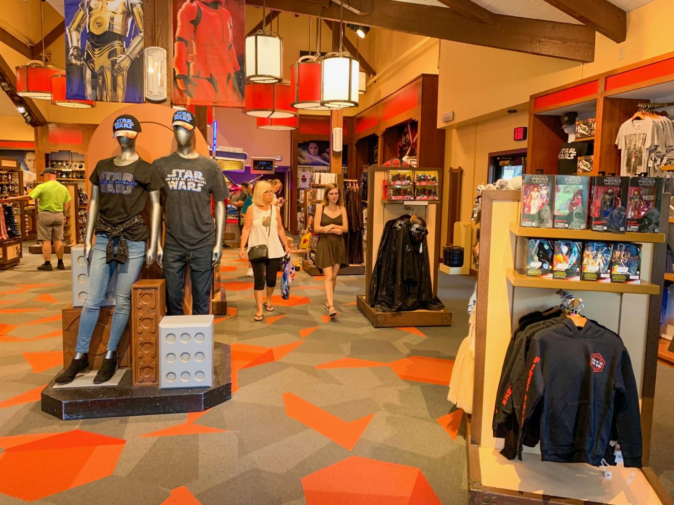 Inside of the Star Wars Trading Post at Disney Springs with large Star Wars artwork on the back wall, tall ceilings, a display in the front right corner with Star Wars clothing, and two mannequins in the center left wearing black Star Wars t shirts, jeans, and black caps with the Star Wars logo.