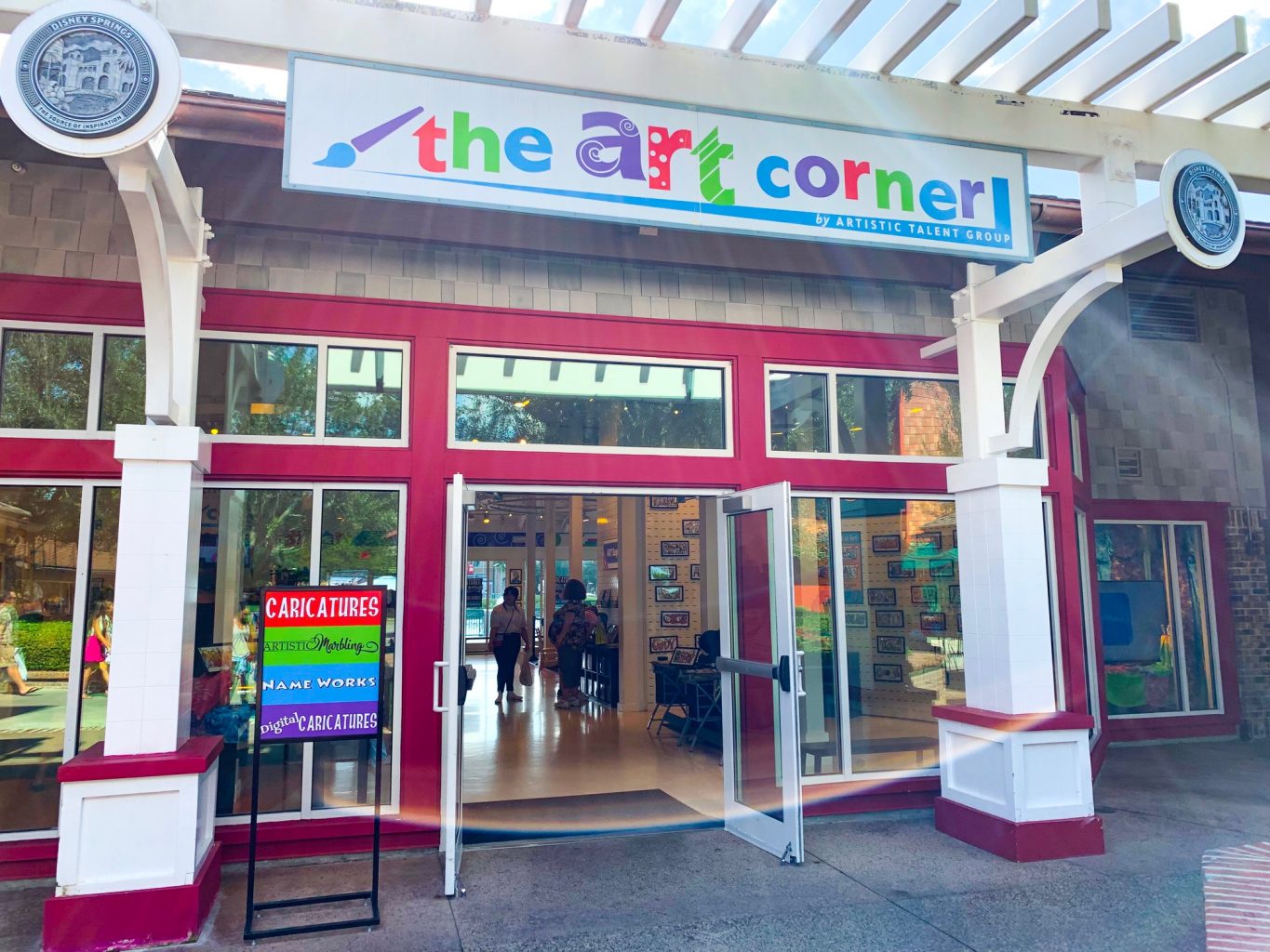 Storefront of The Art Corner at Disney Springs with people inside and red borders around the door and two large windows with a sign above the doors with a white background and a paintbrush with "the art corner" written in multiple colors in a handwriting style font.