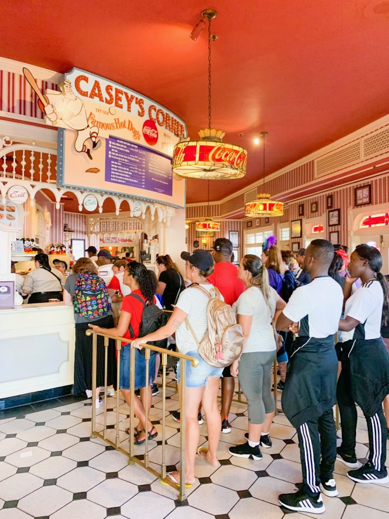 This photo features people standing in a long line to order hot dogs at Casey's, which is one of the best Magic Kingdom restaurants around, and easily found by its  bright red interior and exterior! 