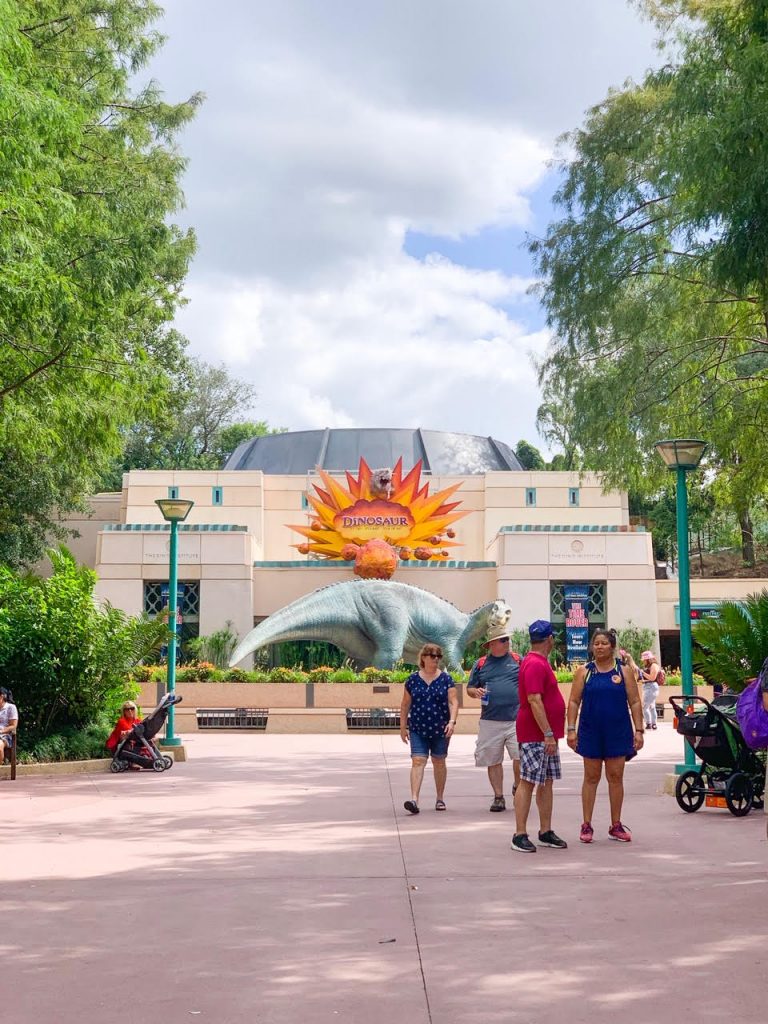 people walking out of building that has large sun and green dinosaur in front, best animal kingdom rides