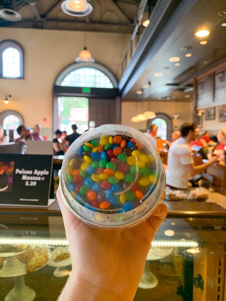 A hand holds up a colorful m&m yogurt parfait in front of a back drop of the inside of Trolley Cafe.