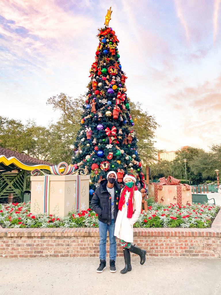 A bundled up couple standing in front of a lavish Christmas tree with giant presents.