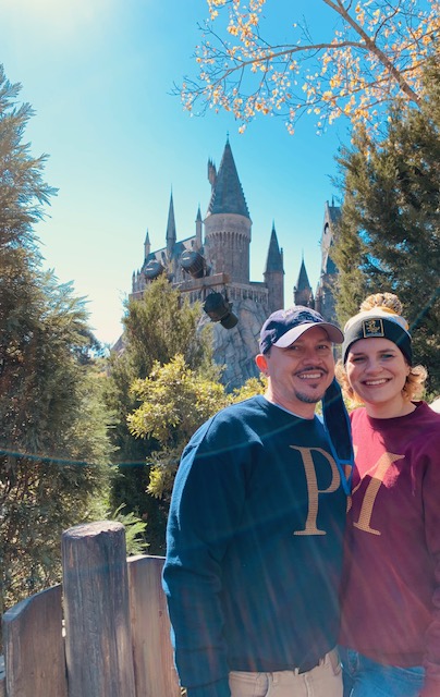 A woman and man in hats and matching sweatshirts stand in front of Hogwarts in the queue of Hagrid. 