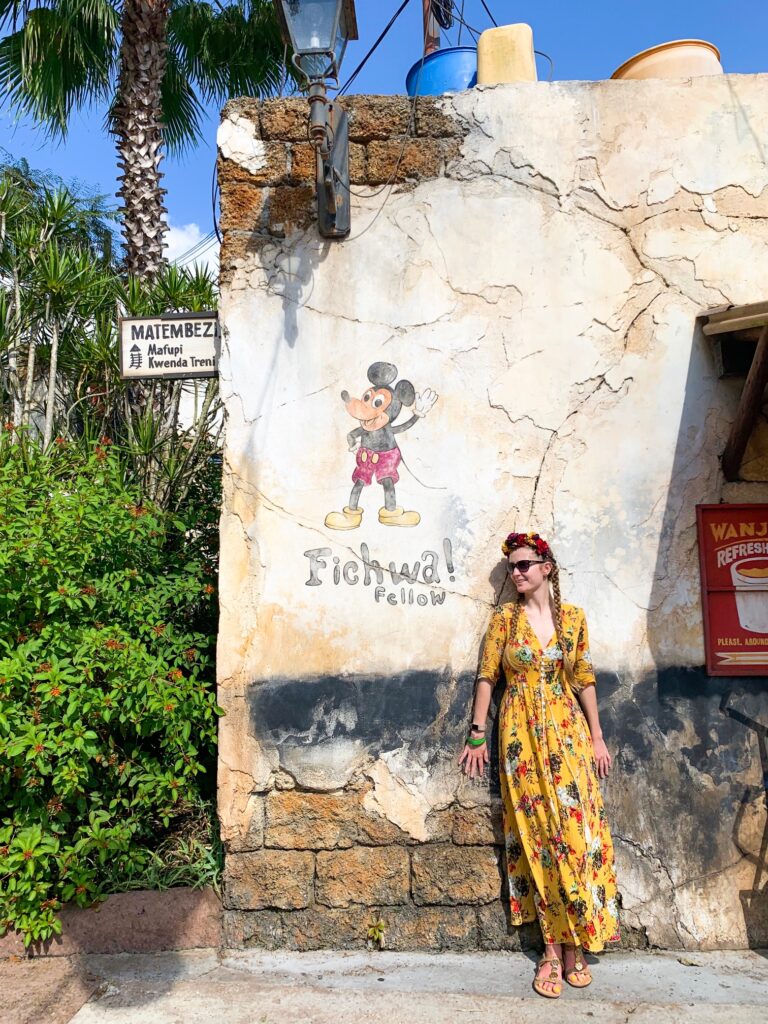 A woman in a yellow, floral dress stands in front of a Mickey Mural, enjoying  Animal Kingdom for adults.
