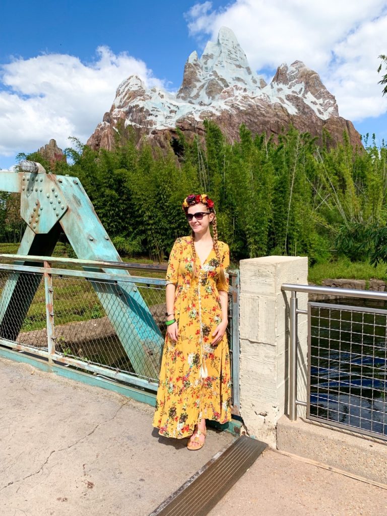 A woman in a yellow, floral dress stands on the bridge in front of the mountain of Expedition Everest. 