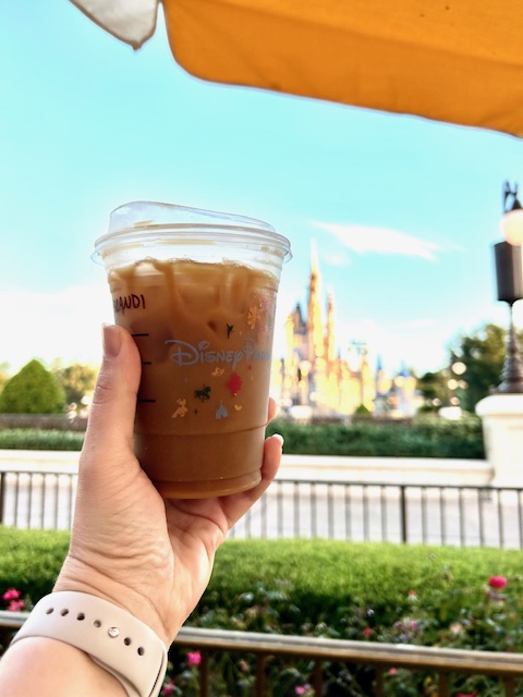 hand holding a Starbucks iced coffee cup in front of blue sky and castle in the background