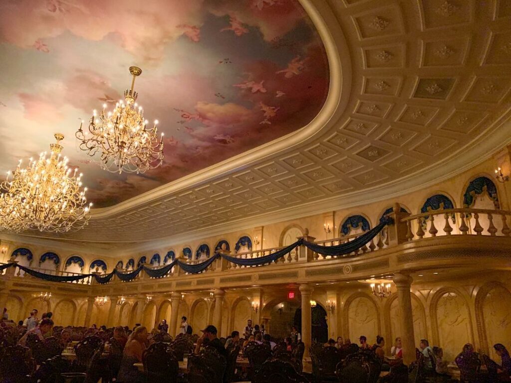 The ceiling is painted with clouds and chandeliers hang over balconies and tables at the Be Our Guest Restaurant. 