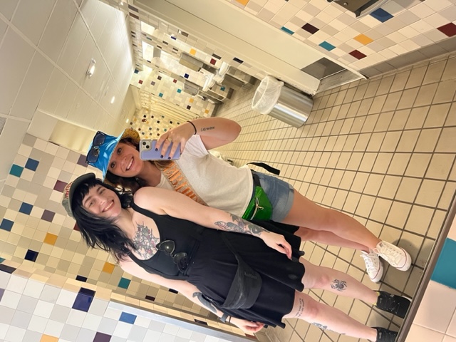 Two girls pose in a bathroom for a mirror selfie, their black and green fanny packs making a splash because they knew not to bring big bags into the park.