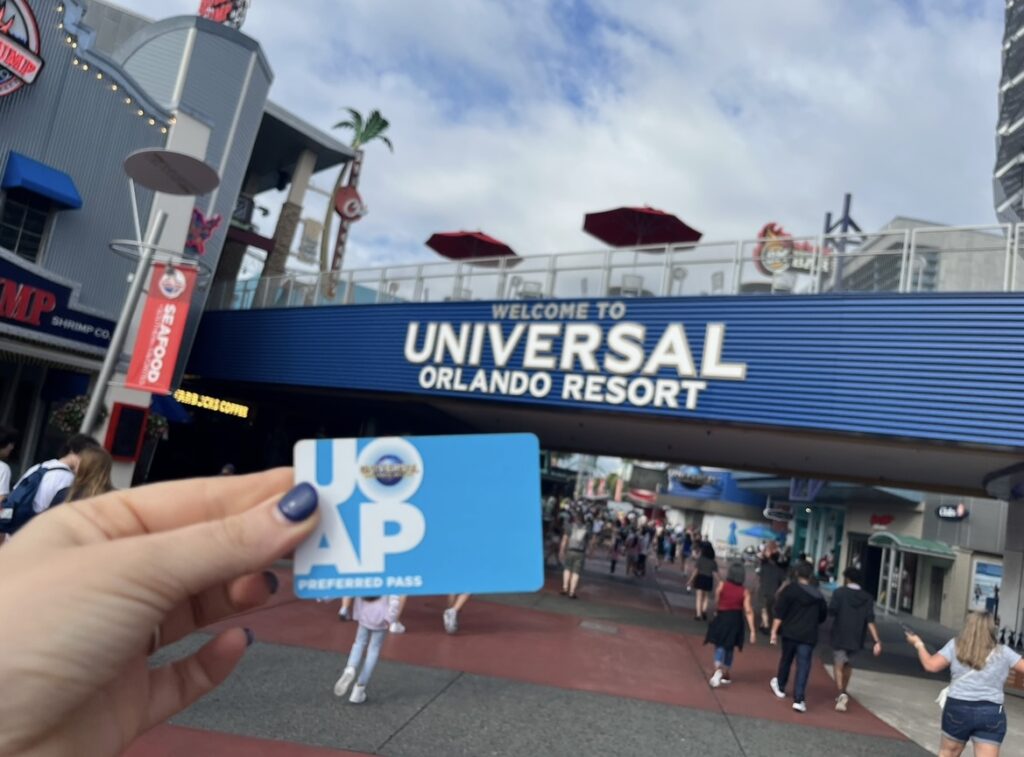 A hand holds up an annual pass, it is blue, and it is the preferred pass. It sits in front of the "Welcome to Universal Orlando Resort" Sign in CityWalk.
