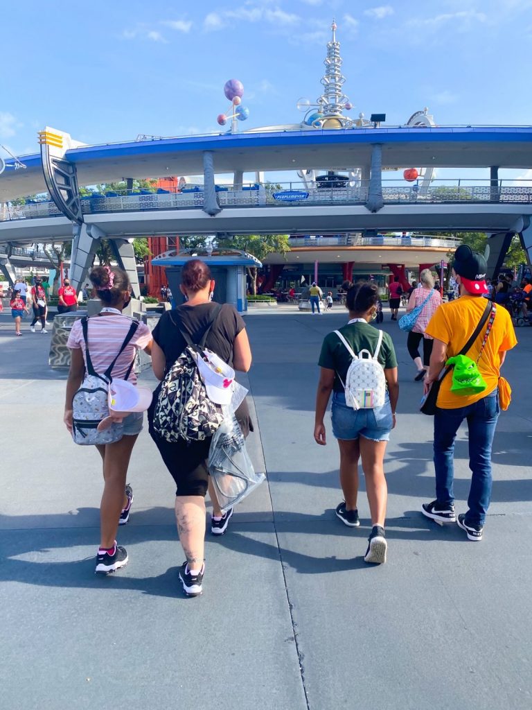 A group of four people walk toward Tomorrowland with backpacks and hats on, obviously glad they prepared the treks by walking more one month before Disney.