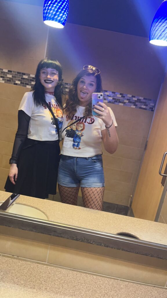 Two girls in Chucky-fan gear stand in a mirror of a bathroom ready to hear Moaning Myrtle, which is one of Universal Orlando Secrets. 