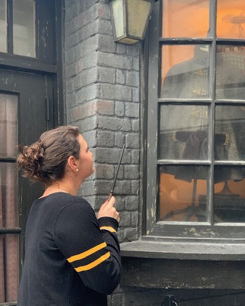 A woman in a hufflepuff sweater points her interactive wand at the glass windows in Diagon Alley. 