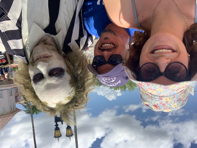 A woman in a bucket hat and a man in a Ravenclaw hat smile with Beetlejuice before a Horror Makeup show. 