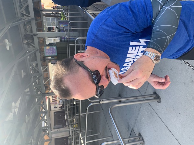 A man in a blue t-shirt and sunglasses eats a beignet as a part of a food sampler which is a great way to save money at Universal Orlando especially during special events.