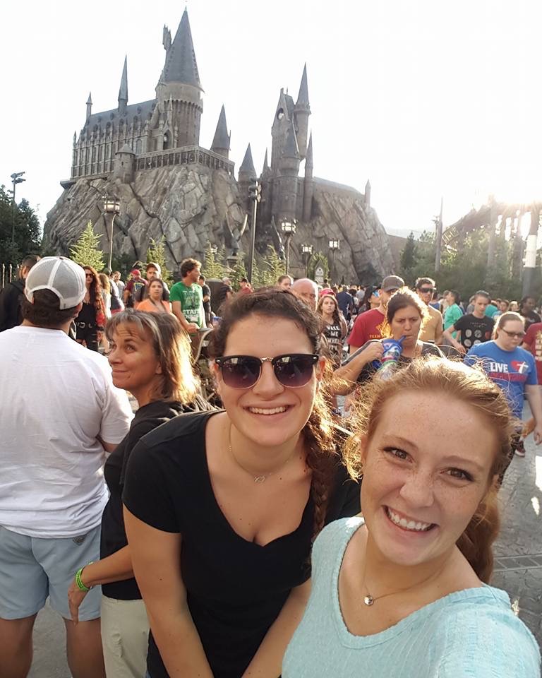 Two girls-- one with red hair and one with a braid-- take a photo in front of Hogwarts during a peak season for the park: the crowd around them is dense. 