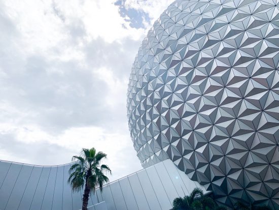 large silver geometric sphere against a cloudy sky and palm tree 