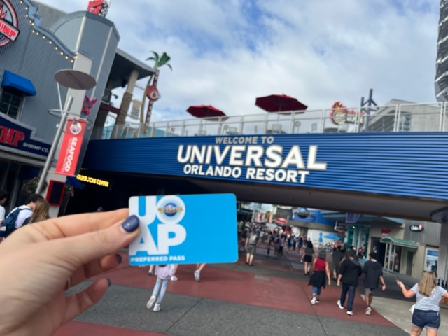 A hand with blue fingernails holds their annual pass up at the "Welcome to Universal Orlando Resort" sign, already wishing they had a lanyard that they bought based off of the list of things to buy from amazon for Universal Studios.