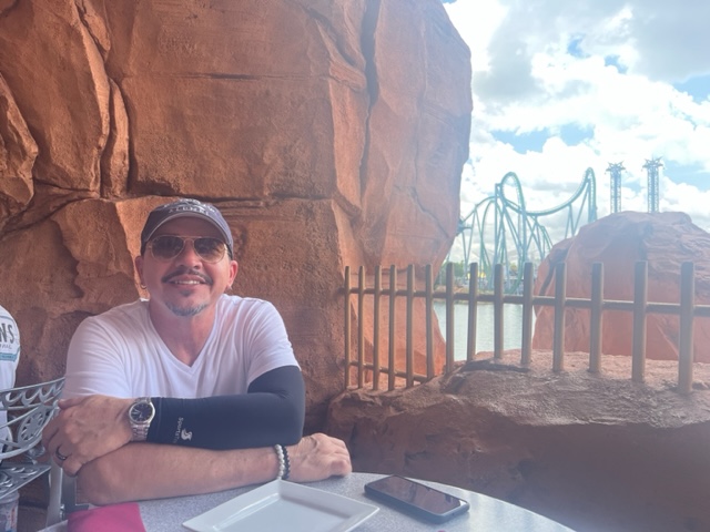 A man in a hat sits in the Mythos restaurant, on the patio, overlooking Dr. Dooms and the Hulk. 