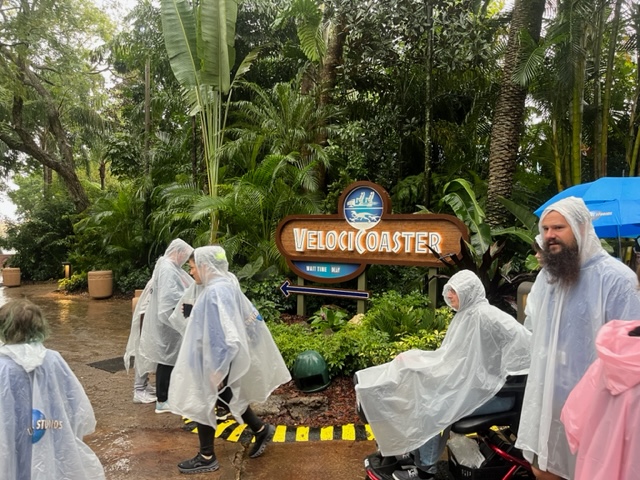 A group of people walk by the Velocicoaster sign in the rain: they are in ponchos, and ponchos are covering wheelchairs and strollers. Invest in stroller covers too! 