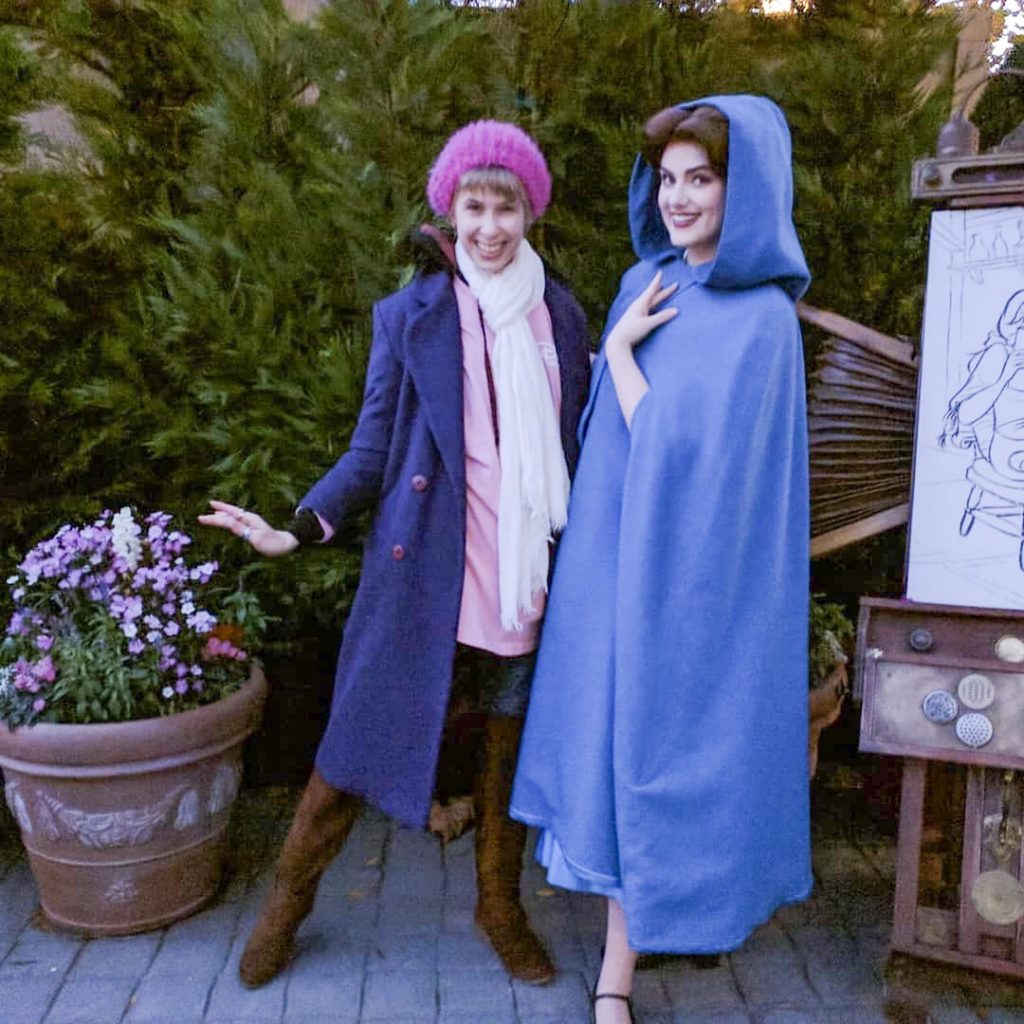 two women in winter clothing, one with a blue cape and the other with a pink hat and blue coat what to wear in disney in winter