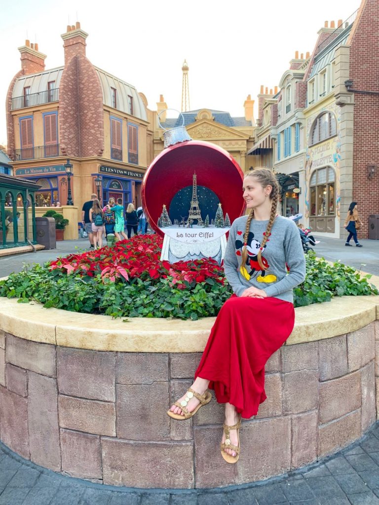 woman in red skirt, sandals, and mickey sweater in front of holiday display and flowers what to wear in disney in winter