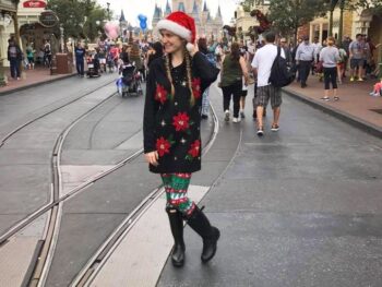 woman in Santa hat, holiday sweater and leggings and boots in front of castle in disney what to wear in disney in winter