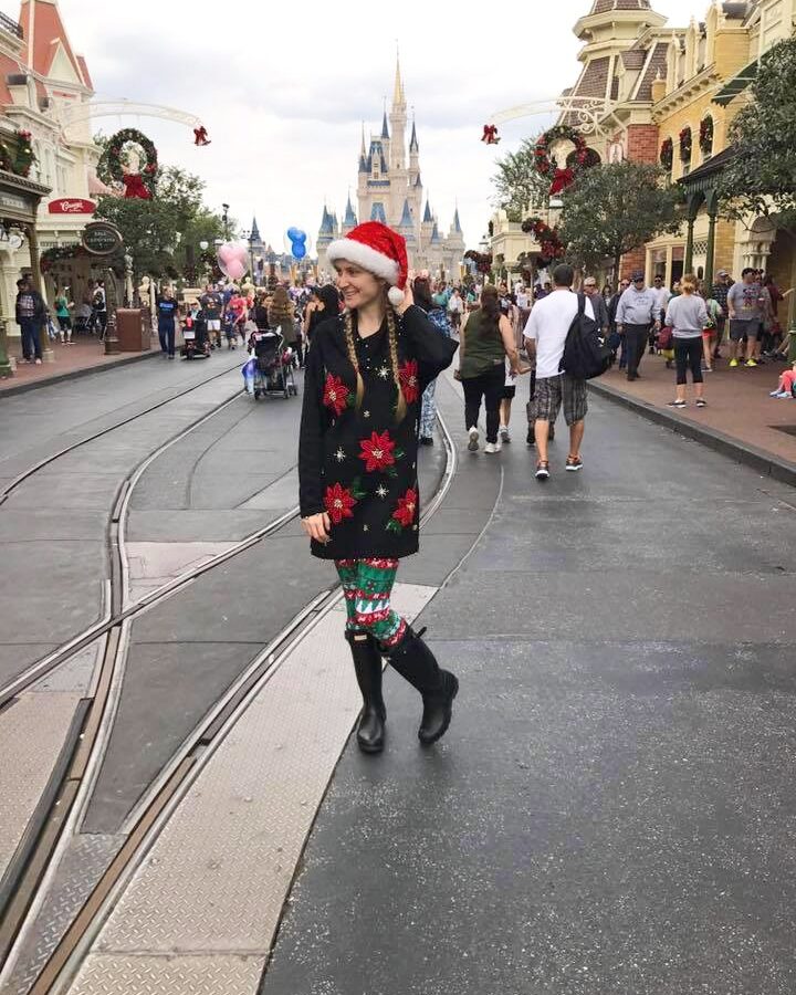 woman in Santa hat, holiday sweater and leggings and boots in front of castle in disney what to wear in disney in winter