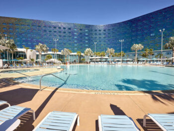 exterior of one of the epic universe hotels at universal orlando. there are loungchairs i the foreground and a pool in the middle and a blue glass hotel in the background. the sky is blue with no clouds. the pavement around the pool is tan