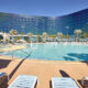 exterior of one of the epic universe hotels at universal orlando. there are loungchairs i the foreground and a pool in the middle and a blue glass hotel in the background. the sky is blue with no clouds. the pavement around the pool is tan