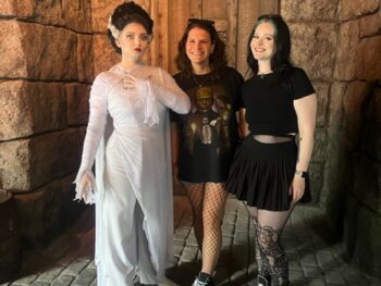 two women in black shorts and tshirts standing next to a employee dressed in costume wearing a white dress and black wig at Epic Universe in Orlando