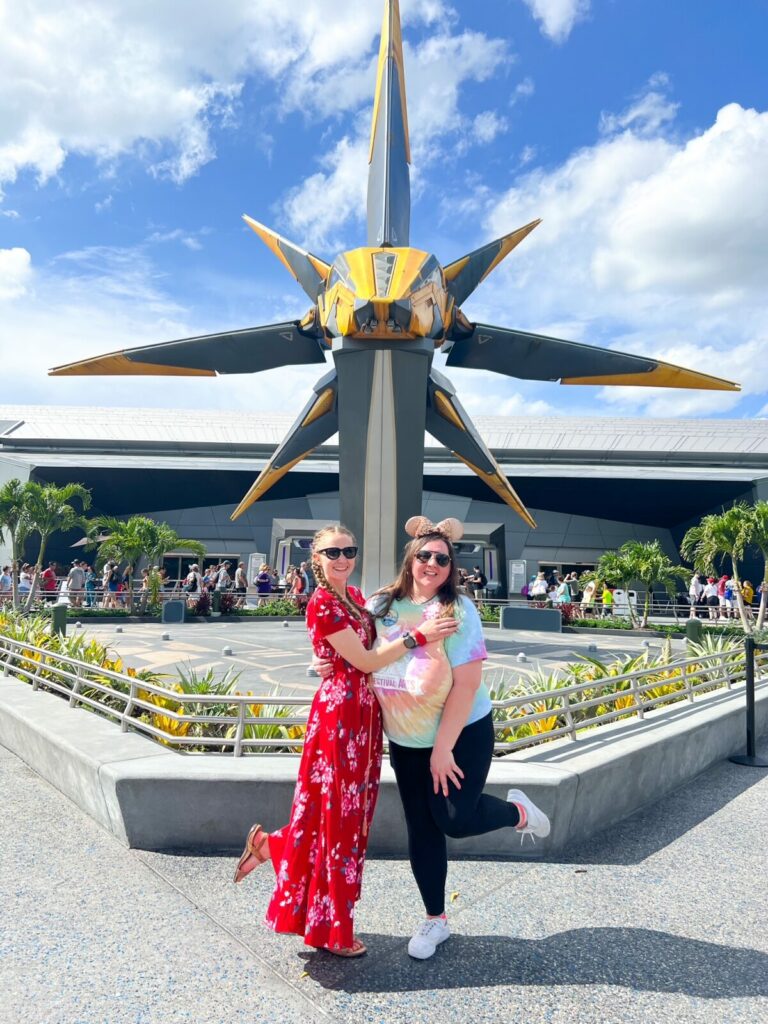 Two sisters, one in a red dress and one in a tie-dye shirt, stand in front of the new Guardians ride at Epcot. 
