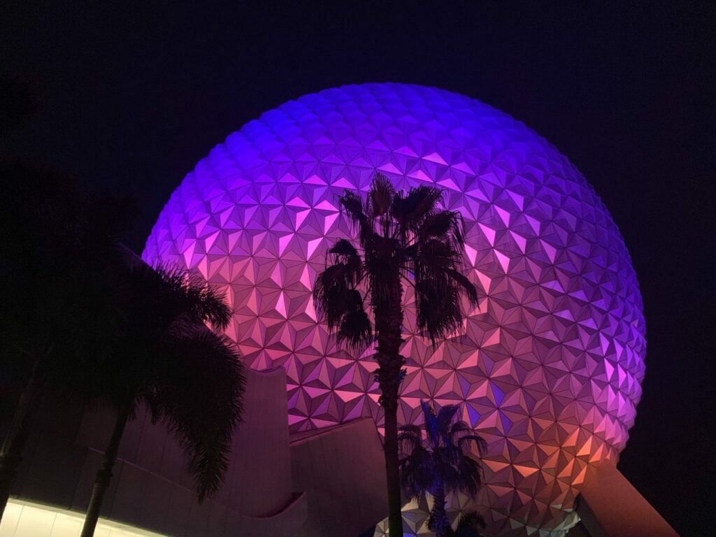 large geodesic sphere lit up purple and pink at night with palm tree in front of it 